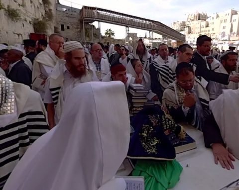 A group of men in traditional Jewish attire standing near a limestone wall, one man touching a table (in Jerusalem, the Holy City)