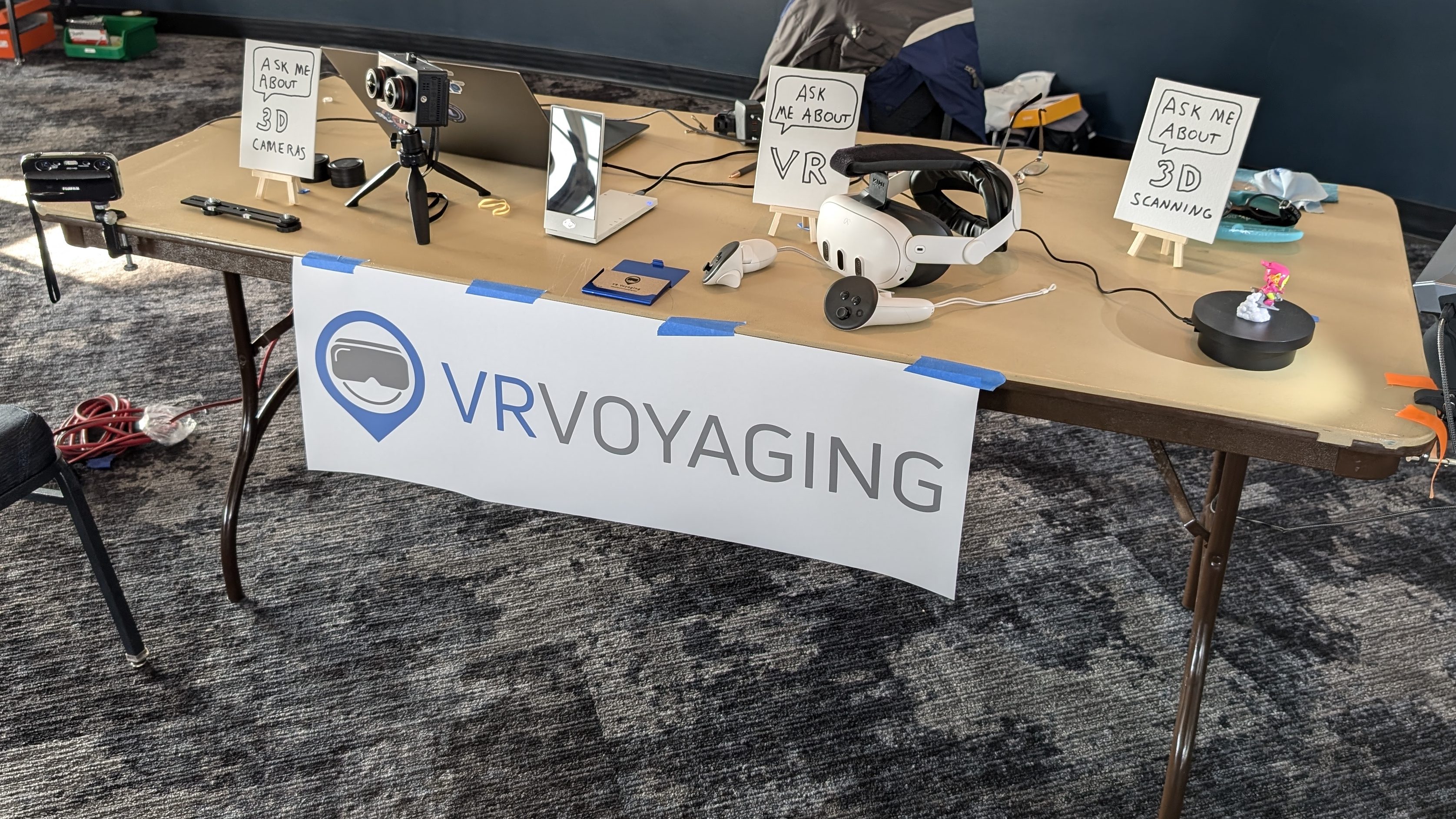 A table with a VR Voyaging banner on the front. Various 3D devices like cameras and a VR headset are set out with labels.